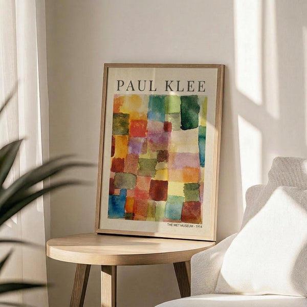 May include: Framed Paul Klee watercolor reproduction featuring colorful geometric squares in vibrant reds, blues, greens, yellows and purples arranged in a grid pattern. The museum poster sits on a round wooden side table next to a beige upholstered chair, with plant leaves casting shadows on white walls in soft natural lighting.