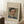 May include: Framed art print featuring a portrait of a man with dark hair and mustache wearing a blue jacket with yellow collar, displayed on white textured rug against white walls with window light, wooden side table with white coffee cup visible nearby