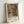 May include: Framed exhibition poster featuring a soft, impressionistic painting of a young woman with long blonde hair wearing a red hat with floral details. The artwork shows gentle brushstrokes in warm tones of pink, green, and coral against a muted background. The poster displays "PIERRE AUGUSTE exhibition" text below the image with "Young Girl with Hat" in smaller text. Light wood frame rests against white wall on white surface, creating subtle shadows.