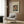 May include: A cream-colored framed poster featuring a vintage portrait of a mustachioed gentleman in blue jacket, displayed on wood paneled wall above a beige upholstered armchair with geometric patterned pillow. Scene includes brass floor lamp with white base and open book on dark round side table near window with curtains.