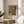 May include: Framed poster featuring a soft impressionist painting of a young woman with blonde hair wearing a pink ribbon hat, displayed on a round wooden side table next to a beige upholstered chair. Natural sunlight streams across white walls, with green plant leaves visible in the corner. The artwork shows delicate brushstrokes in muted greens, pinks, and cream tones within a thin gold frame.