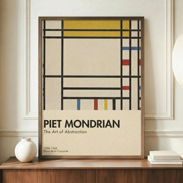 May include: Framed abstract art poster with geometric grid pattern in black lines, featuring yellow, blue, and red rectangular sections on cream background. Exhibition-style text reads "PIET MONDRIAN The Art of Abstraction 1938-1943 Place de la Concorde" below the artwork. Displayed on wooden sideboard next to white ceramic vase and stacked books in elegant room with cream walls and white molding.