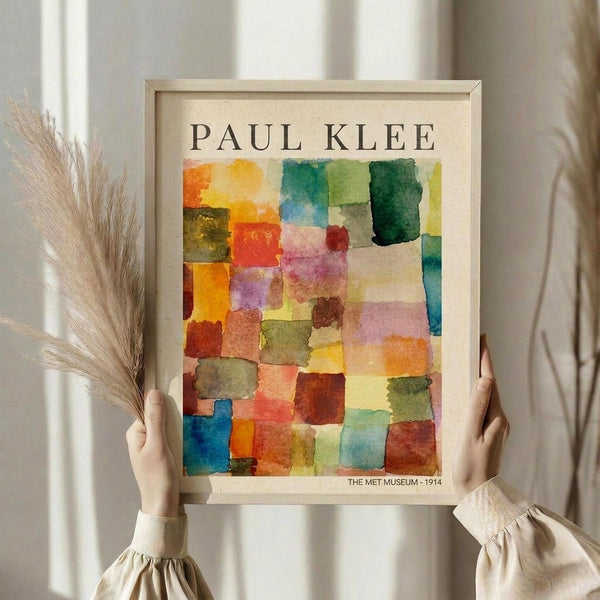 May include: Hands holding framed Paul Klee abstract watercolor print featuring colorful geometric squares in warm oranges, reds, yellows, cool blues and greens. Light wooden frame against cream background with dried pampas grass stems visible to left. Person wearing beige ruffled sleeve top displaying museum poster reproduction.