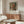 May include: Framed art print of a young woman wearing an ornate floral hat displayed above a mid-century modern walnut credenza with brass hardware. Room features cream walls, white upholstered chair, glass-top oval coffee table, and brass floor lamp. Print shows impressionist-style painting in soft greens, pinks, and earth tones with exhibition text below the image.