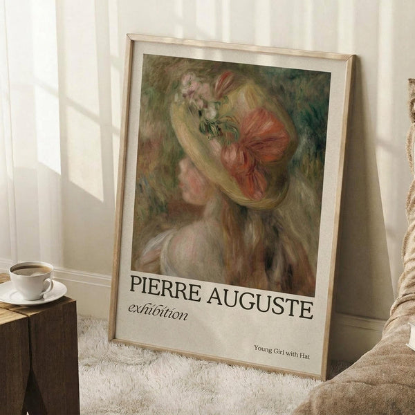 May include: A framed exhibition poster featuring a soft, impressionistic painting of a young woman with flowing blonde hair and a red bow or flower accent, displayed in a light wooden frame leaning against white paneled walls. The poster has cream-colored text at the bottom. Scene includes a white coffee cup on a wooden block and textured beige throw blanket, creating a cozy interior setting with natural window lighting.