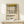 May include: Framed poster displaying Piet Mondrian's geometric abstract art with black grid lines, yellow rectangle at top, small red and blue rectangles on right side, and cream background. Text reads "PIET MONDRIAN The Art of Abstraction 1938-1943 Place de la Concorde" in black letters. Light wood frame sits on white wooden surface next to small wrapped gift with twine and greenery against cream wall.