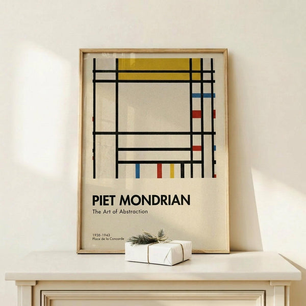 May include: Framed poster displaying Piet Mondrian's geometric abstract art with black grid lines, yellow rectangle at top, small red and blue rectangles on right side, and cream background. Text reads "PIET MONDRIAN The Art of Abstraction 1938-1943 Place de la Concorde" in black letters. Light wood frame sits on white wooden surface next to small wrapped gift with twine and greenery against cream wall.