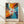 May include: Framed abstract expressionist poster with explosive radiating design in vibrant blues, oranges, and reds emanating from white center with red circle. Dynamic paint splatters and streaks create energetic burst pattern. Natural wood frame displays against white wall on light wooden floor with diagonal shadows.
