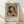 May include: Framed art print leaning against white wall on light wood floor. Shows Renoir's impressionist painting of young girl in pink feathered bonnet and rose-colored dress, hand posed near chin. Exhibition poster format with "PIERRE AUGUSTE" text and artwork details below image. Natural wood frame with light matting. Soft window light creates diagonal shadows on textured white wall.