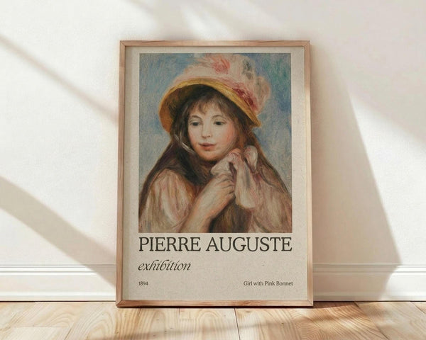 May include: Framed art print leaning against white wall on light wood floor. Shows Renoir's impressionist painting of young girl in pink feathered bonnet and rose-colored dress, hand posed near chin. Exhibition poster format with "PIERRE AUGUSTE" text and artwork details below image. Natural wood frame with light matting. Soft window light creates diagonal shadows on textured white wall.