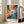 May include: Framed abstract expressionist poster featuring explosive radiating brushstrokes in vibrant orange, yellow, red, and blue with scattered dots and splatters emanating from a central white form with red circle. Text reads "POLLOCK Expressive Expressionist Movement" below artwork. Light wood frame leaning against white wall next to beige sofa in bright, sunlit room with large windows.