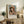 May include: Framed art print featuring a young girl wearing a pink and white bonnet, displayed on a rustic wooden sideboard in a cozy living room setting. The poster shows warm peachy tones and soft brushstrokes in an impressionist style. Scene includes cream-colored furniture, potted fern, ceramic vase, lamp, and stacked books creating a warm, inviting home decor atmosphere.
