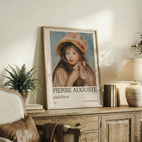 May include: Framed art print featuring a young girl wearing a pink and white bonnet, displayed on a rustic wooden sideboard in a cozy living room setting. The poster shows warm peachy tones and soft brushstrokes in an impressionist style. Scene includes cream-colored furniture, potted fern, ceramic vase, lamp, and stacked books creating a warm, inviting home decor atmosphere.