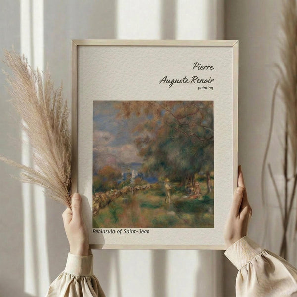 May include: Person in cream-colored clothing holding framed reproduction of Renoir's impressionist landscape painting showing pastoral scene with figures, trees, and soft brushstrokes in greens, blues, and earth tones. Light wooden frame with cream matting displays artwork title and artist name. Dried pampas grass visible beside frame against neutral curtained background.