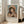 May include: Framed poster print leaning against white wall next to beige sofa in bright living room. Artwork shows young girl in yellow hat with pink feathers, holding small dog, painted in soft impressionist style with warm tones. Exhibition poster format with "PIERRE AUGUSTE" text and "1894" date below painting. Natural sunlight streams through windows onto light wooden floors.
