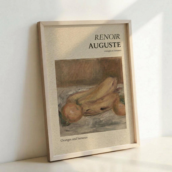 May include: A framed art print featuring Auguste Renoir's painting of oranges and bananas in warm yellow and orange tones against a muted brown background. The print has cream-colored matting with "RENOIR AUGUSTE" and "oranges et bananes" text at the top, and "Oranges and bananas" at the bottom. The natural wood frame leans against a white wall, casting soft shadows on a light surface.