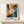 May include: Abstract expressionist artwork poster in light wood frame on white shelf beside small wrapped gift. Painting features explosive radial composition with orange, red, yellow, blue, and black paint streaks radiating from white center with red circle. Dynamic splashes, drips, and circular dots create energetic movement. White wall background, minimal modern interior setting.