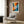 May include: Modern living room with beige armchair and geometric pillow, featuring a framed abstract art poster on wood-paneled wall. The artwork displays dynamic radiating brushstrokes in vibrant orange, blue, and black with a central white spiral containing a red dot. Gold floor lamp and black side table with open book complete the contemporary interior scene.