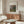 May include: Framed Renoir landscape print hanging above a wooden sideboard in a modern living room. The artwork shows an impressionist countryside scene with rolling hills, trees, and soft brushstrokes in greens, blues, and earth tones. White matting surrounds the print in a thin wooden frame. Room features cream walls, white curved chair, glass oval coffee table, and minimalist white table lamp.