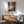 May include: Abstract expressionist poster print in wooden frame featuring explosive radiating brushstrokes in vibrant blues, oranges, and reds with white center and red circle accent. Displayed above mid-century modern wooden credenza with white lamp, next to sage green upholstered armchair in bright, naturally lit room with white walls.