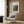 May include: Framed Renoir landscape print displayed on wood paneled wall above beige upholstered chair with geometric pillow. Artwork shows impressionist painting with soft brushstrokes depicting trees and figures in muted greens, blues, and earth tones. Modern reading nook with brass floor lamp and side table.