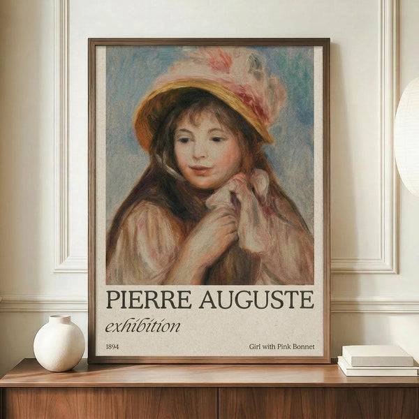 May include: Framed poster print featuring a soft impressionist portrait of a young girl in a yellow straw bonnet with pink ribbon, her hands positioned near her face wearing pale clothing. Exhibition-style poster with "PIERRE AUGUSTE" and "exhibition" text below the artwork, dated 1894 with "Girl with Pink Bonnet" caption. Displayed on warm wood sideboard next to small white ceramic vase and stacked books against cream paneled wall.