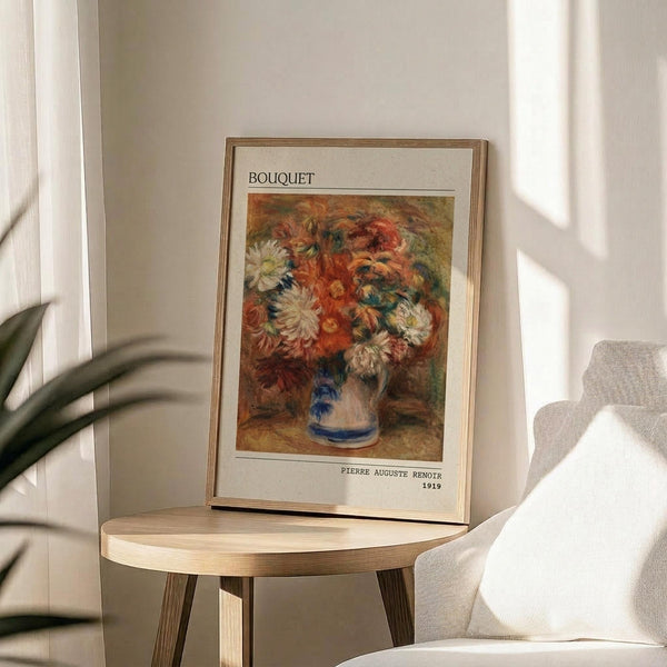 May include: Framed art print of Renoir's 1919 floral painting featuring vibrant orange, white and red flowers in a blue and white ceramic vase, displayed on a light wood round side table next to a beige upholstered chair. Soft natural lighting creates gentle shadows on white walls with plant leaves visible in corner.