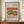 May include: Framed art print displaying Renoir's painting of a green pear and red apple on a beige background, positioned on a wooden sideboard next to a white ceramic vase and books, with a woven pendant light visible in an elegant room setting with cream walls and decorative molding.