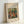 May include: Framed art print leaning against white wall showing Renoir's impressionist painting of people in a pastoral scene with warm colors of orange, green, and yellow. The wooden frame surrounds a cream-colored mat with black text identifying the artist and artwork title. Soft natural lighting creates gentle shadows on the wall behind the rectangular frame.