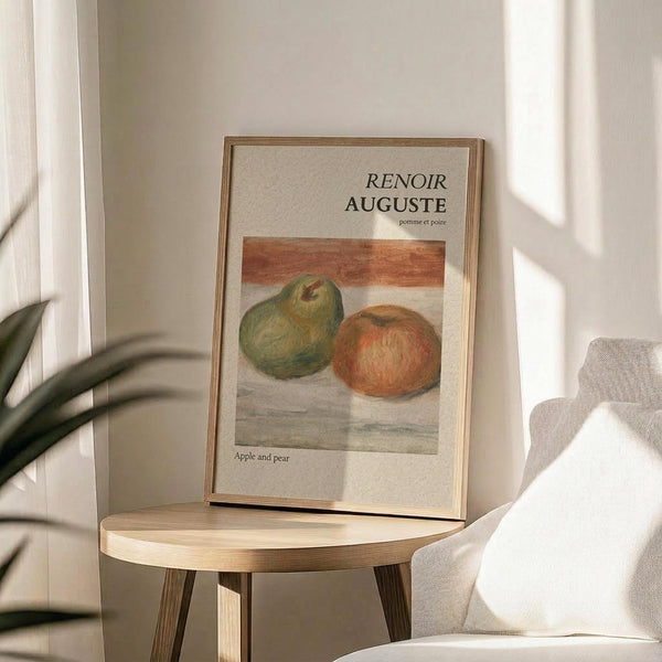 May include: Framed art print displaying Renoir's still life painting of green pear and red apple on neutral background, positioned on light wood round side table next to beige upholstered chair, with plant leaves visible in soft natural lighting against white walls.