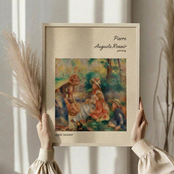 May include: Hands holding a framed reproduction of Renoir's impressionist painting showing figures in a pastoral scene with warm oranges, greens, and earth tones. The artwork is displayed in a light wood frame with cream matting against soft white curtains, alongside dried wheat stalks for styling.