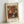 May include: Framed art print leaning against white wall showing impressionist painting of colorful flower bouquet in blue and white striped vase. Artwork features vibrant orange, red, white and yellow blooms with loose brushstrokes in warm, golden tones. Print has white border with "BOUQUET" at top and "PIERRE AUGUSTE RENOIR 1919" at bottom, displayed in light wooden frame.