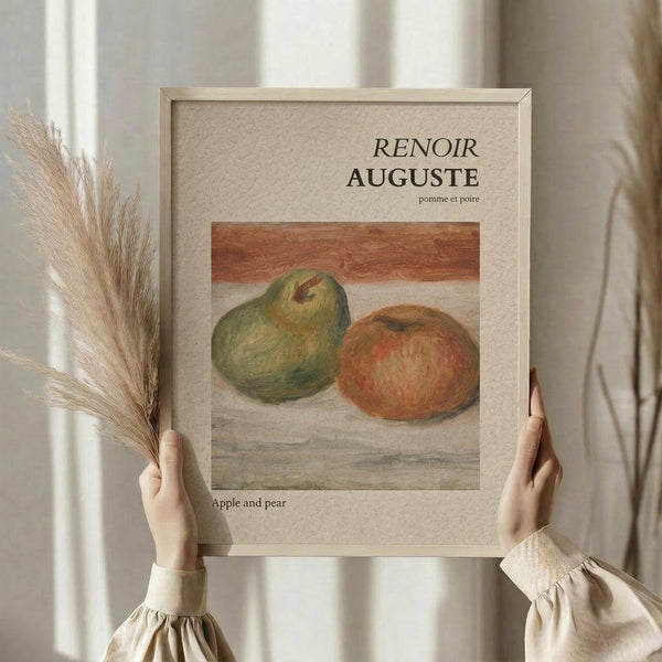 May include: Person in cream sweater holding framed art print showing Renoir's still life painting of a green pear and red apple on neutral surface. Light wood frame with beige matting. Dried pampas grass visible on left side. Soft natural lighting against white curtained background creates minimalist styling scene.