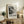 May include: Black and white abstract art poster in light wood frame, displayed on rustic wooden sideboard with fern plant, books, and ceramic vase. The artwork features splattered paint textures and organic shapes in monochrome tones, leaning against white wall in cozy living room setting with beige upholstered furniture and warm natural lighting.