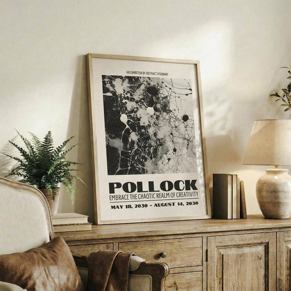 May include: Black and white abstract art poster in light wood frame, displayed on rustic wooden sideboard with fern plant, books, and ceramic vase. The artwork features splattered paint textures and organic shapes in monochrome tones, leaning against white wall in cozy living room setting with beige upholstered furniture and warm natural lighting.