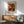 May include: Framed art print of Renoir's colorful floral bouquet painting from 1919, featuring vibrant orange, red, and white flowers in a blue and white ceramic vase. The poster hangs above a mid-century modern wooden credenza with a white table lamp. A sage green upholstered armchair with wooden frame sits nearby, with warm sunlight casting geometric shadows across the white wall and wooden floor.