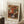 May include: Framed art print of an Impressionist floral painting leaning against white wainscoting wall. Features vibrant bouquet with orange, red, and white flowers in blue-striped ceramic vase, painted in loose brushstrokes. Cream matting with "BOUQUET" at top and "PIERRE AUGUSTE RENOIR 1919" at bottom. Wooden side table with white coffee cup and saucer visible on left, beige textured rug below, cozy home interior setting.