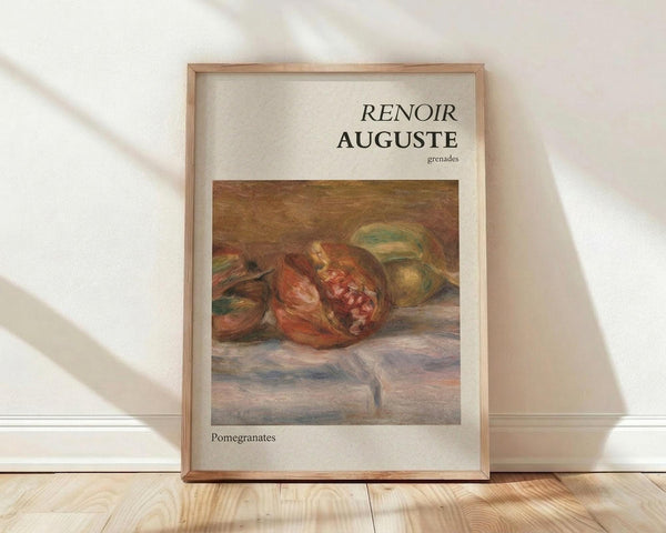 May include: Framed art print displaying Renoir's impressionist still life painting of pomegranates in warm reddish-brown tones with green fruit, mounted in light wood frame against cream textured wall on wooden floor with soft natural lighting and shadows