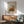 May include: Framed poster of autumn trees with orange and golden foliage in impressionist painting style, displayed above modern wood credenza with white ceramic table lamp. Mid-century sage green armchair with wooden arms positioned nearby in bright, minimalist room with white walls and natural sunlight creating geometric shadows.
