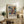 May include: Framed abstract art poster with vibrant blue, orange, yellow and red brushstrokes displayed on rustic wooden sideboard. Room scene shows white walls, fern plant in ceramic pot, stacked books, clay vase, and brown leather chair. Natural lighting creates soft shadows. Poster features exhibition-style text below colorful abstract painting in light wood frame.