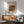 May include: Framed art print featuring Auguste Renoir's still life painting of a green striped melon and peaches in warm earth tones, displayed above a mid-century modern wooden sideboard with white ceramic lamp. The poster has cream matting in a thin wood frame, positioned in a minimalist room setting with sage green upholstered armchair, round coffee table, and natural sunlight casting geometric shadows on white walls.