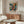 May include: Framed abstract art poster with vibrant blue, orange, and yellow paint splatters in Jackson Pollock style, displayed above mid-century walnut credenza in modern living room. White curved chair and glass coffee table visible. Poster features bold "POLLOCK" text and exhibition dates. Warm natural lighting creates shadows on cream walls.