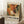 May include: Framed abstract art poster with vibrant orange, green, and cream paint splatters in Jackson Pollock style, leaning against white wall near wooden side table with coffee cup. Features exhibition-style text reading "POLLOCK - EMBRACE THE CHAOTIC REALM OF CREATIVITY" with dates "MAY 18, 2030 - AUGUST 14, 2030" and "AN EXHIBITION BY ABSTRACT VISIONARY" at top. Soft lighting creates shadows on textured rug beside beige fabric.