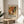 May include: Framed exhibition poster featuring abstract splattered artwork in vibrant oranges, greens, and reds with white splatters, displayed on round wooden side table next to beige upholstered chair. Natural sunlight streams across neutral wall creating soft shadows. Green plant leaves visible in corner of minimalist modern living space.