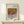 May include: Framed art print displaying Renoir's impressionist still life painting of ripe pomegranates in warm red and orange tones against muted background, positioned on light wood console table beside small white wrapped gift with evergreen sprig, against cream colored wall with natural lighting