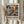 May include: Person holding framed abstract art poster featuring vibrant swirls of blue, orange, yellow, and coral colors in chaotic brushstroke patterns. Text reads "AN EXHIBITION BY ABSTRACT VISIONARY" at top and "POLLOCK" in large letters below artwork, followed by "EMBRACE THE CHAOTIC REALM OF CREATIVITY" and exhibition dates. Light wooden frame against neutral curtained background with dried pampas grass visible on left side.