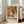 May include: Large framed poster with abstract paint splatter artwork in vibrant oranges, greens, and earth tones, featuring white paint splatters and organic flowing shapes. Exhibition-style design with "POLLOCK" text and creative tagline below the artwork. Light wood frame leaning against white wall beside beige upholstered chair in bright, modern living room with large windows.