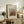 May include: Framed art print featuring Pierre Auguste Renoir's impressionist painting of autumn trees in warm oranges, reds and greens against a soft blue sky. The beige matted artwork is displayed on a rustic wooden sideboard alongside books, a ceramic vase, and a fern plant. A cream upholstered chair with brown throw pillows sits nearby in this cozy, neutral-toned living space with white walls.