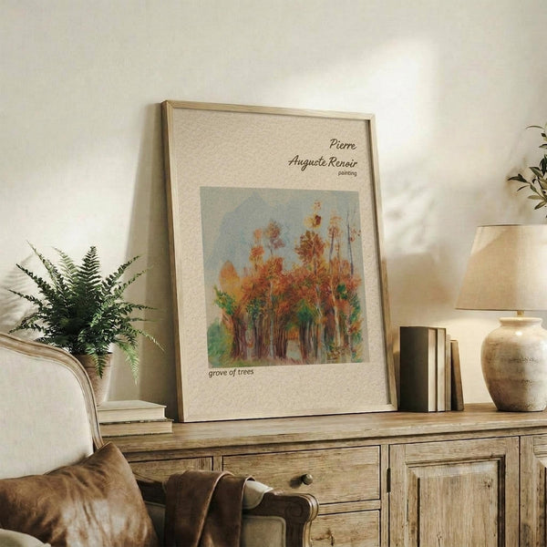 May include: Framed art print featuring Pierre Auguste Renoir's impressionist painting of autumn trees in warm oranges, reds and greens against a soft blue sky. The beige matted artwork is displayed on a rustic wooden sideboard alongside books, a ceramic vase, and a fern plant. A cream upholstered chair with brown throw pillows sits nearby in this cozy, neutral-toned living space with white walls.