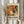 May include: Person holding framed abstract art poster featuring vibrant orange, red, green and cream paint splatters in chaotic dripping style. Poster displays exhibition text for Pollock show dates May-August 2030. Light wood frame against white curtained background with dried wheat stalks visible on left side.